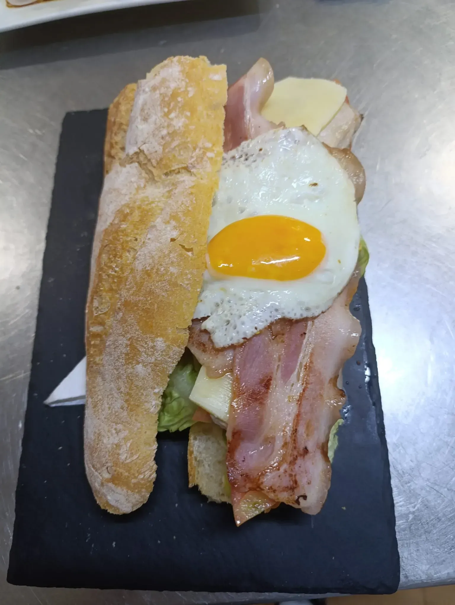 Sándwich en bandeja negra: baguette, huevo frito, tocino, queso, lechuga.