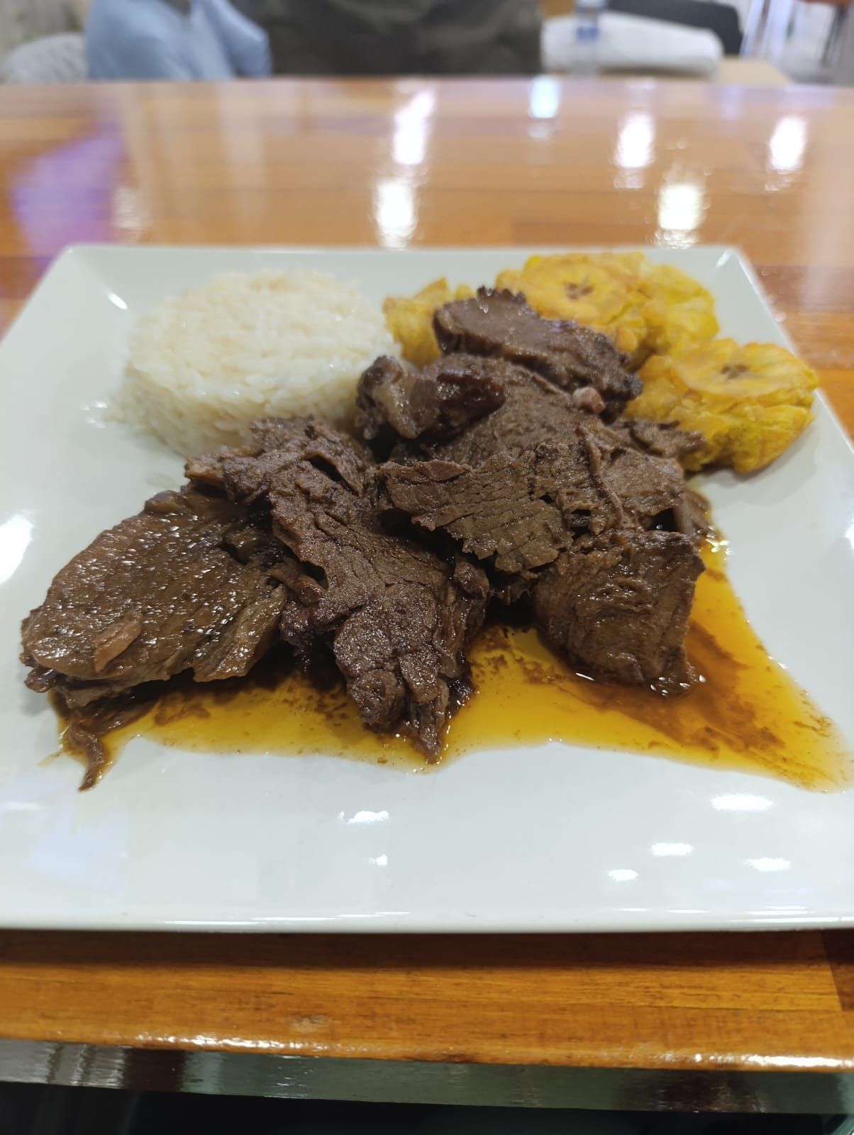 Plato de carne cocida con arroz y plátanos fritos en una mesa de madera.