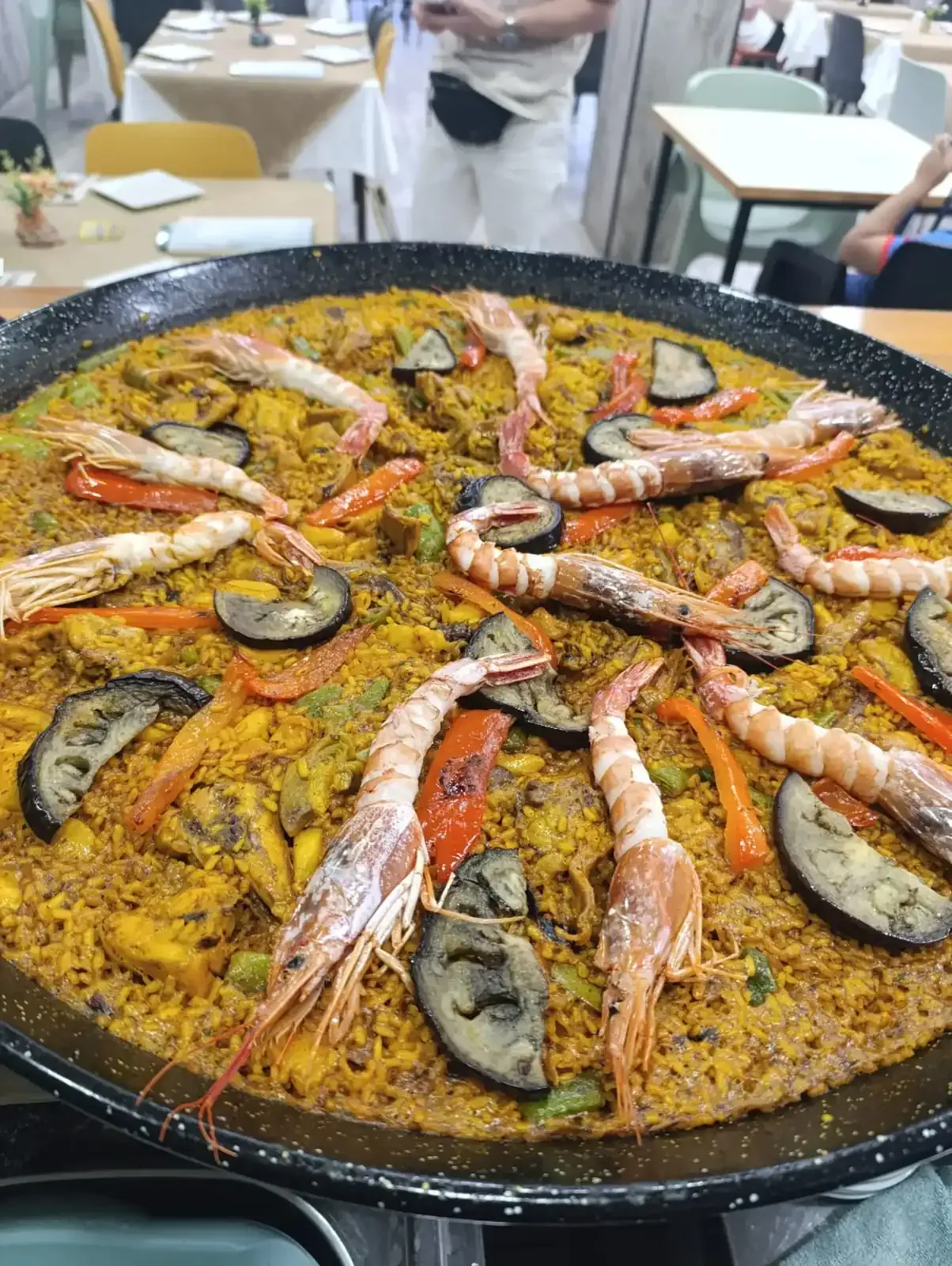 Paellera grande con camarones, verduras y arroz amarillo, en ambiente de restaurante.