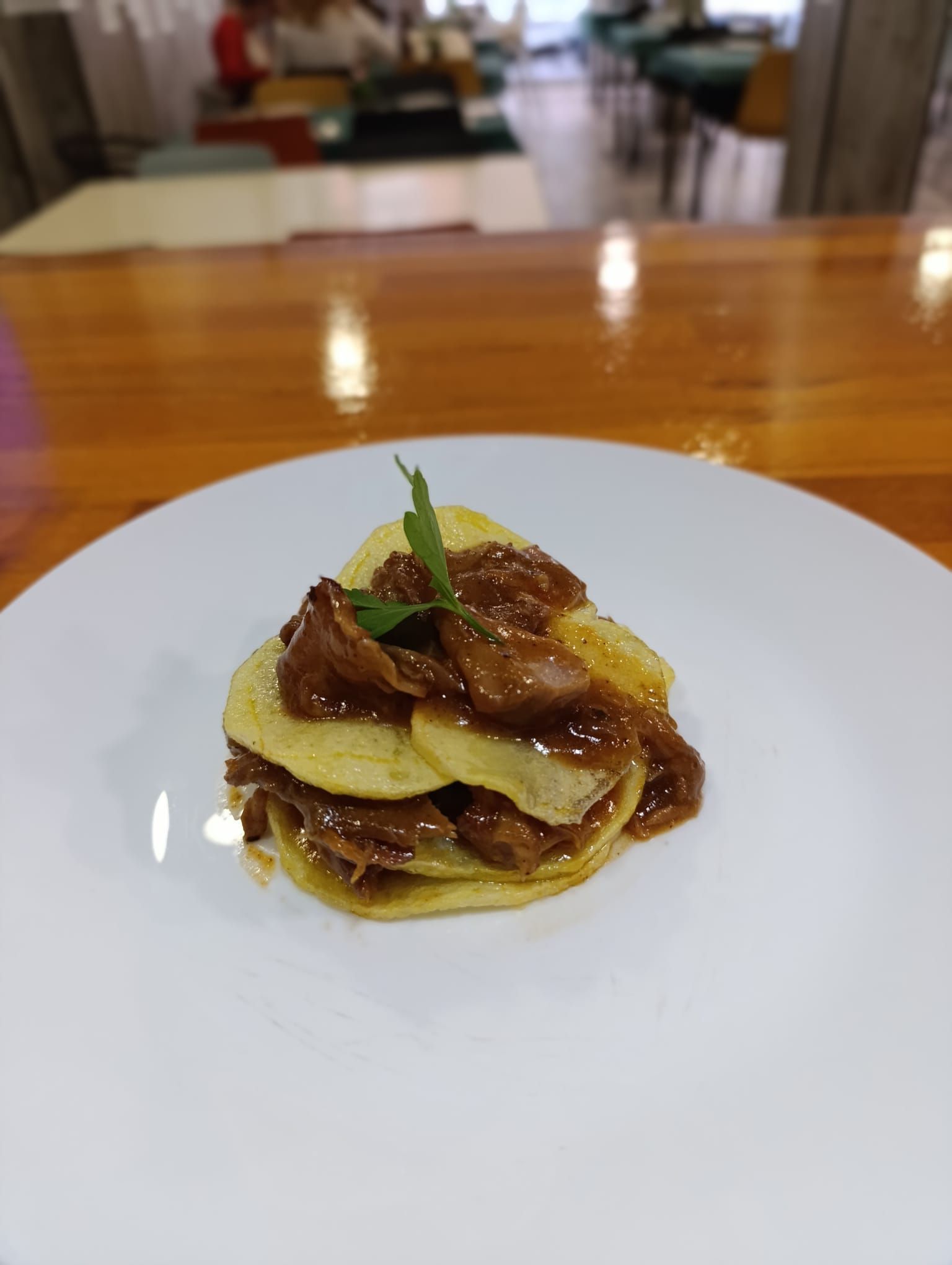 Rebanadas de papa apiladas cubiertas con carne oscura y salsa en un plato blanco.