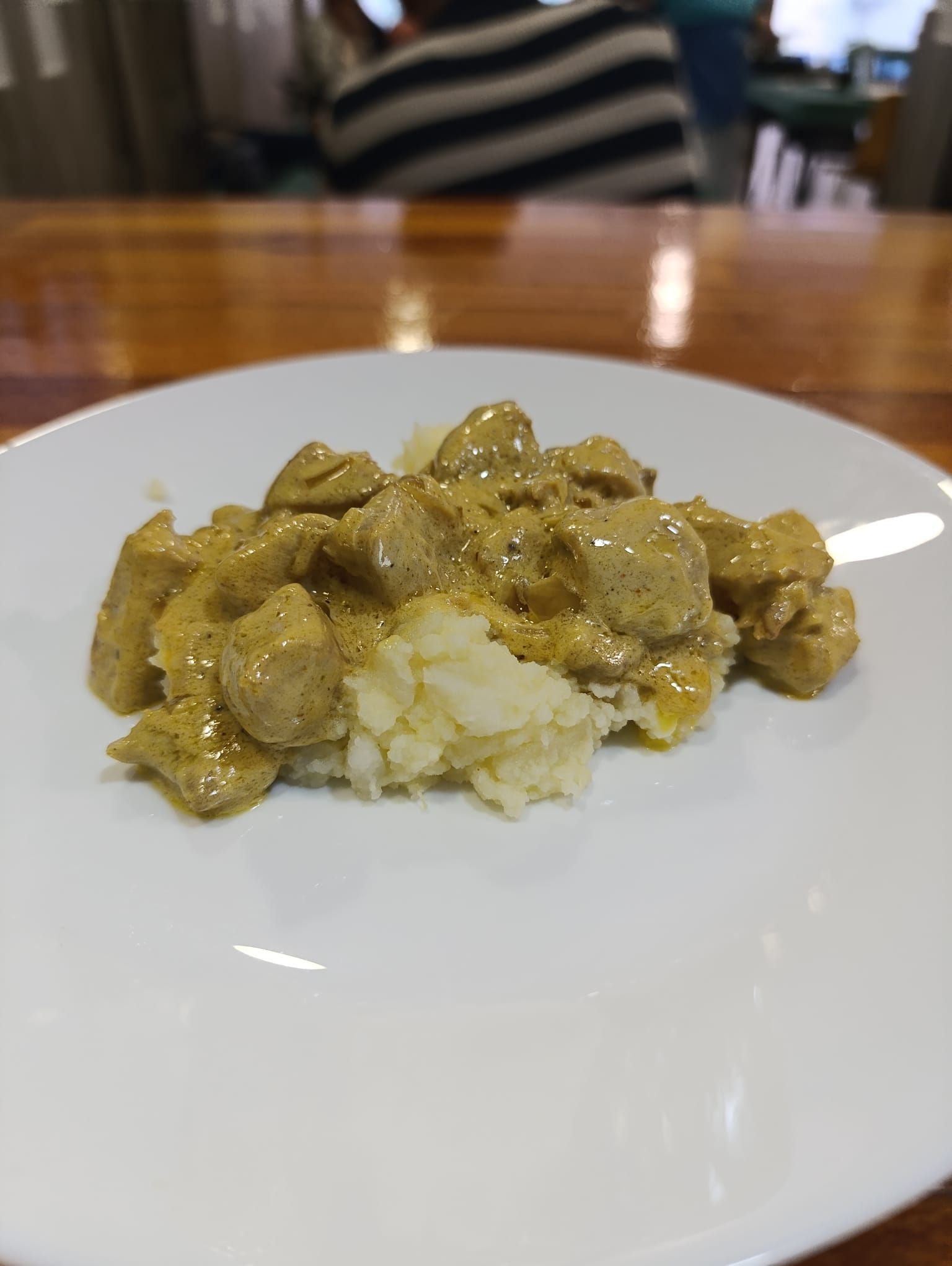 Pollo al curry sobre puré de patatas en un plato blanco, mesa de madera al fondo.
