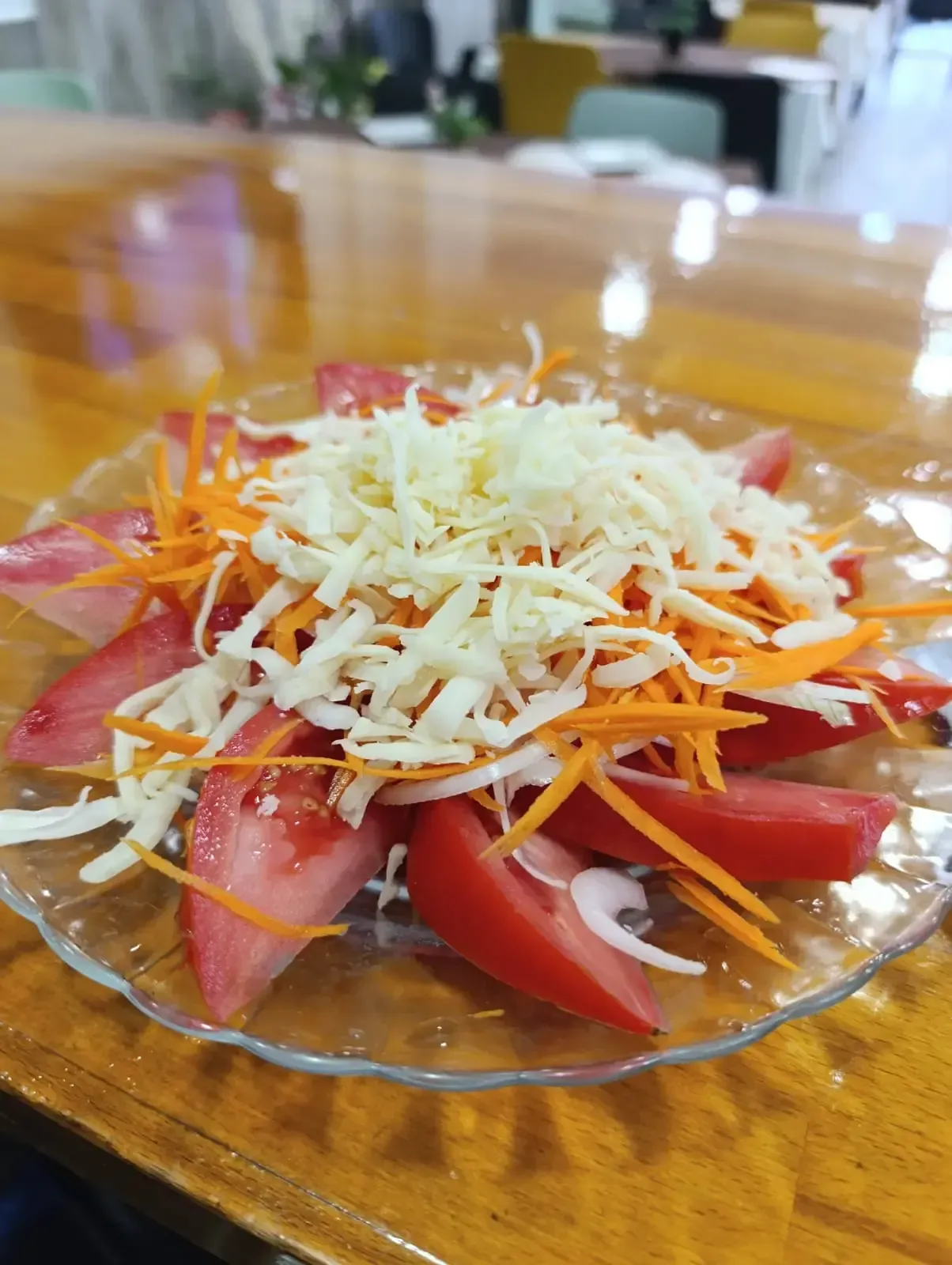 Ensalada de tomate con zanahorias ralladas y queso en un plato transparente, sobre una mesa de madera.