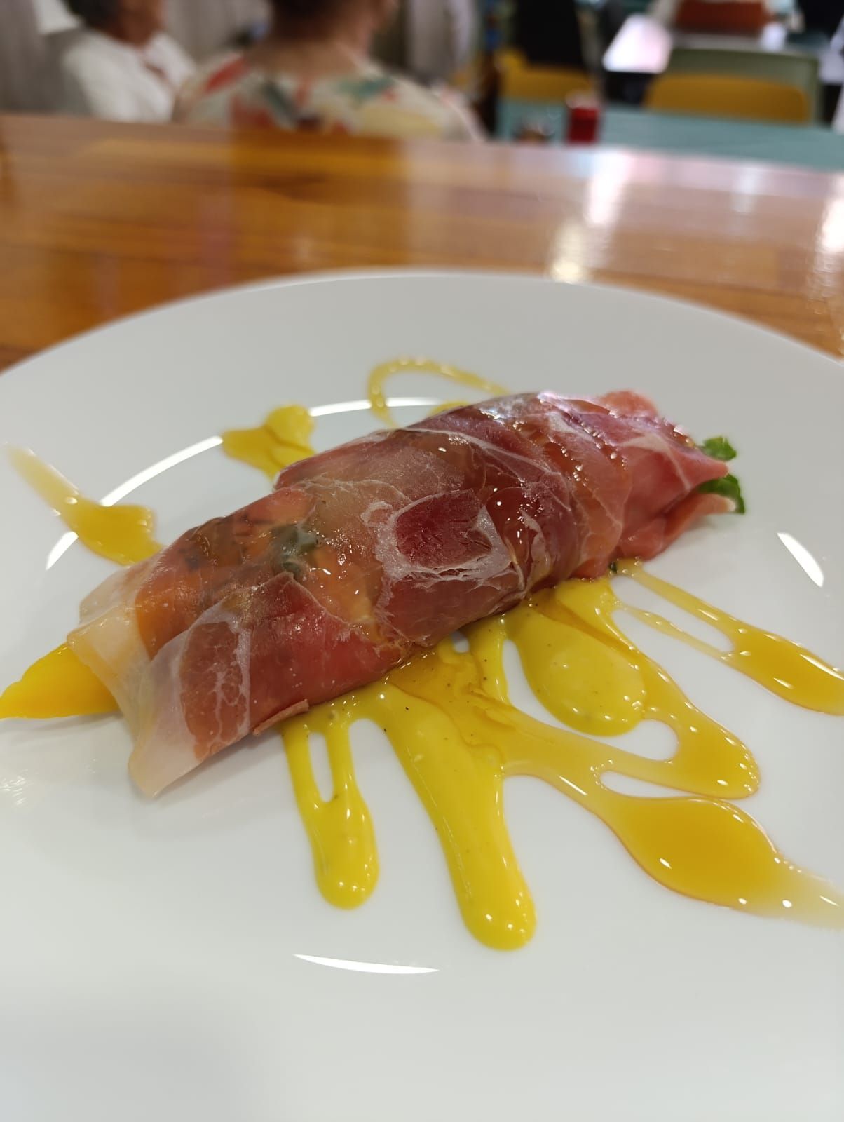 Comida envuelta en prosciutto, con un toque de salsa amarilla, en un plato blanco. En un restaurante.