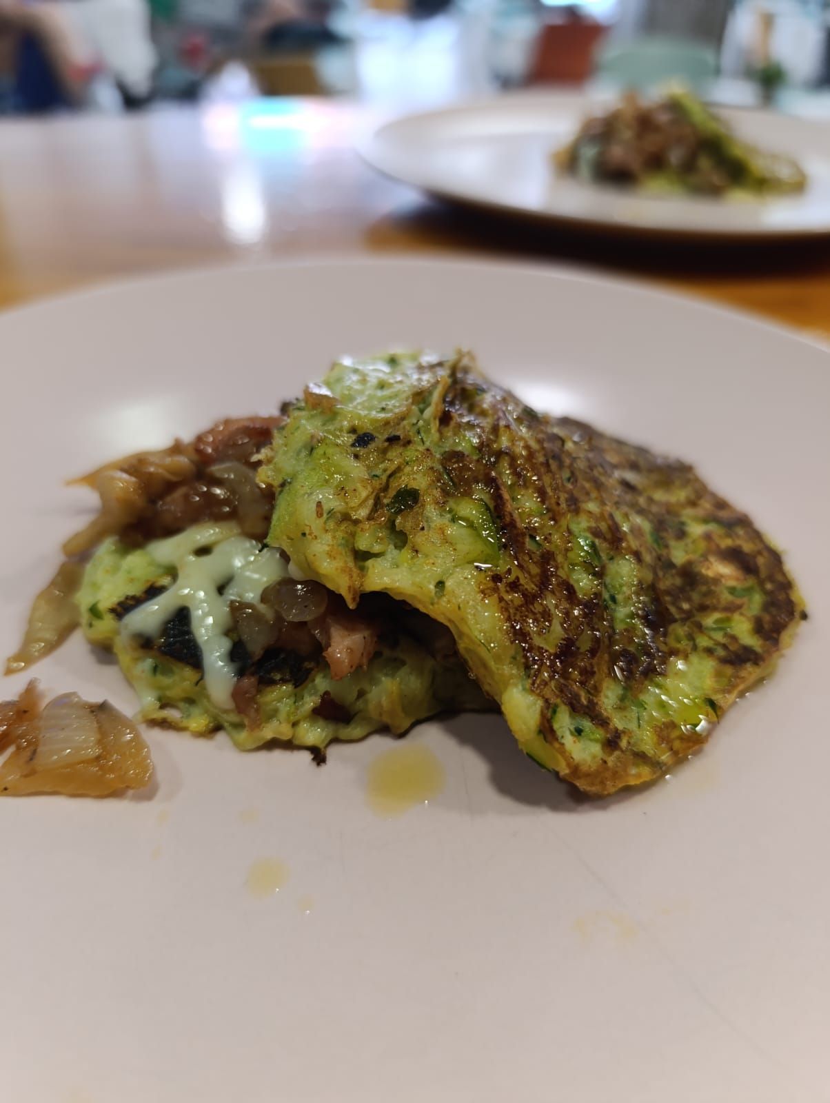 Dos panqueques verdes fritos en un plato blanco, cubiertos con cebollas caramelizadas y un chorrito.