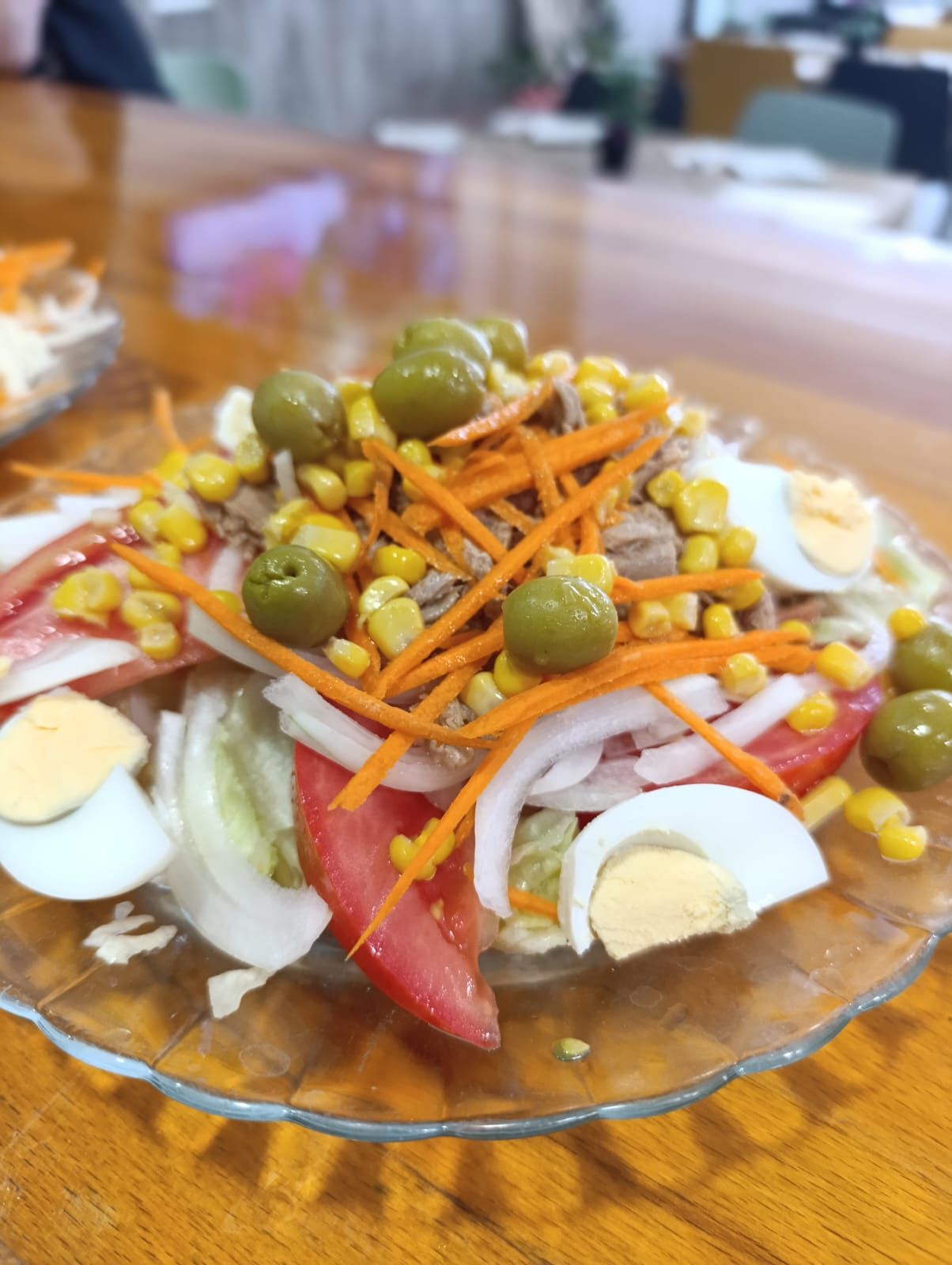 Ensalada con aceitunas, maíz, zanahorias, tomates en rodajas, cebollas, huevos y lechuga en un plato de vidrio.