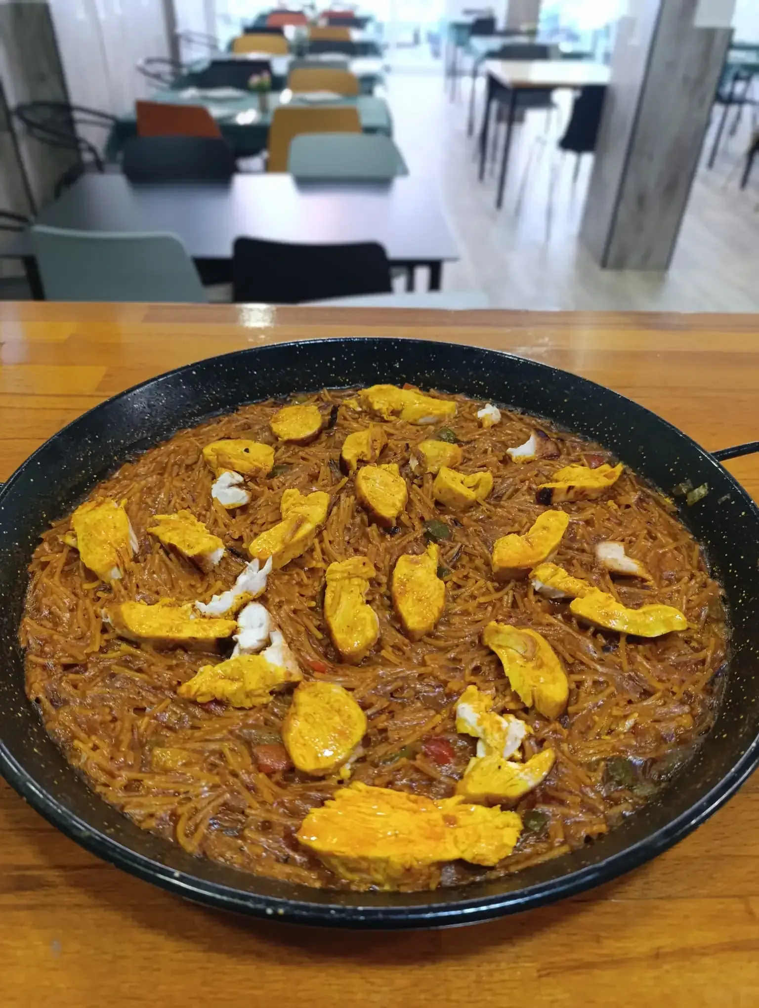 Una sartén de fideuá (paella de fideos) con trozos de pescado amarillo sobre una mesa de madera, en un entorno de cafetería.