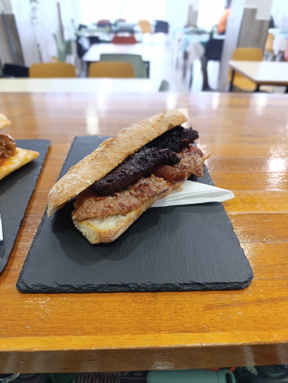 Sándwich sobre pizarra oscura, con carne y salchicha oscura en su interior, sobre una mesa de madera.