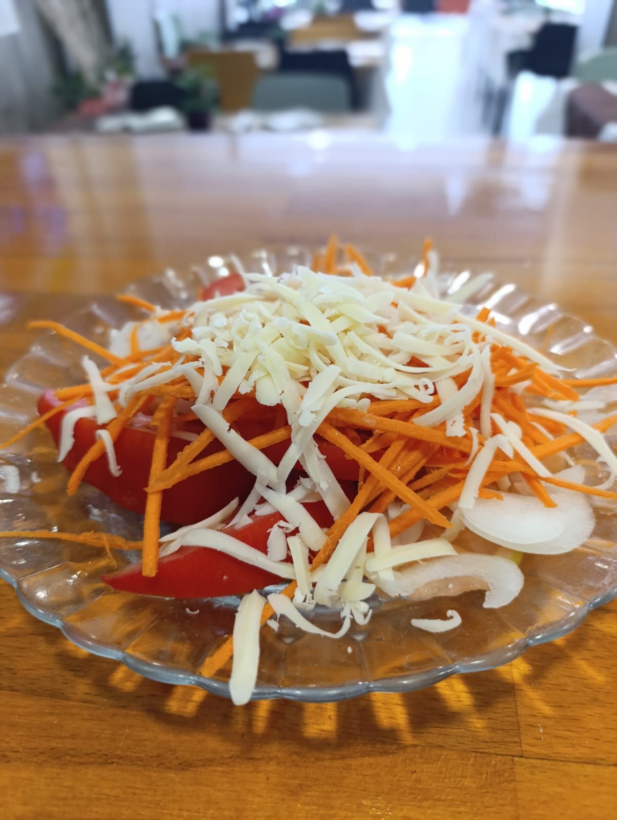 Ensalada en un plato: tomates en rodajas, zanahorias, cebolla, cubierto con queso rallado, sobre una mesa de madera.