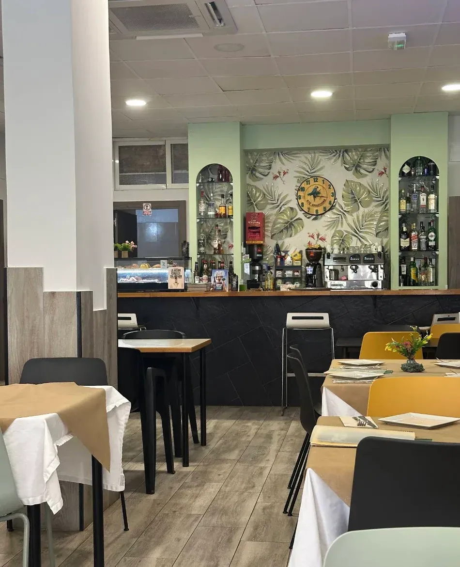 Interior de un restaurante con mesas, barra y papel pintado botánico, iluminado por luces cenitales.