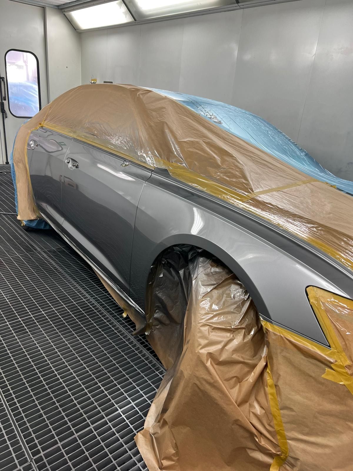 Seitenansicht eines hellgrauen Autos in der Lackierkabine mit abgeklebten Scheiben und Dach.