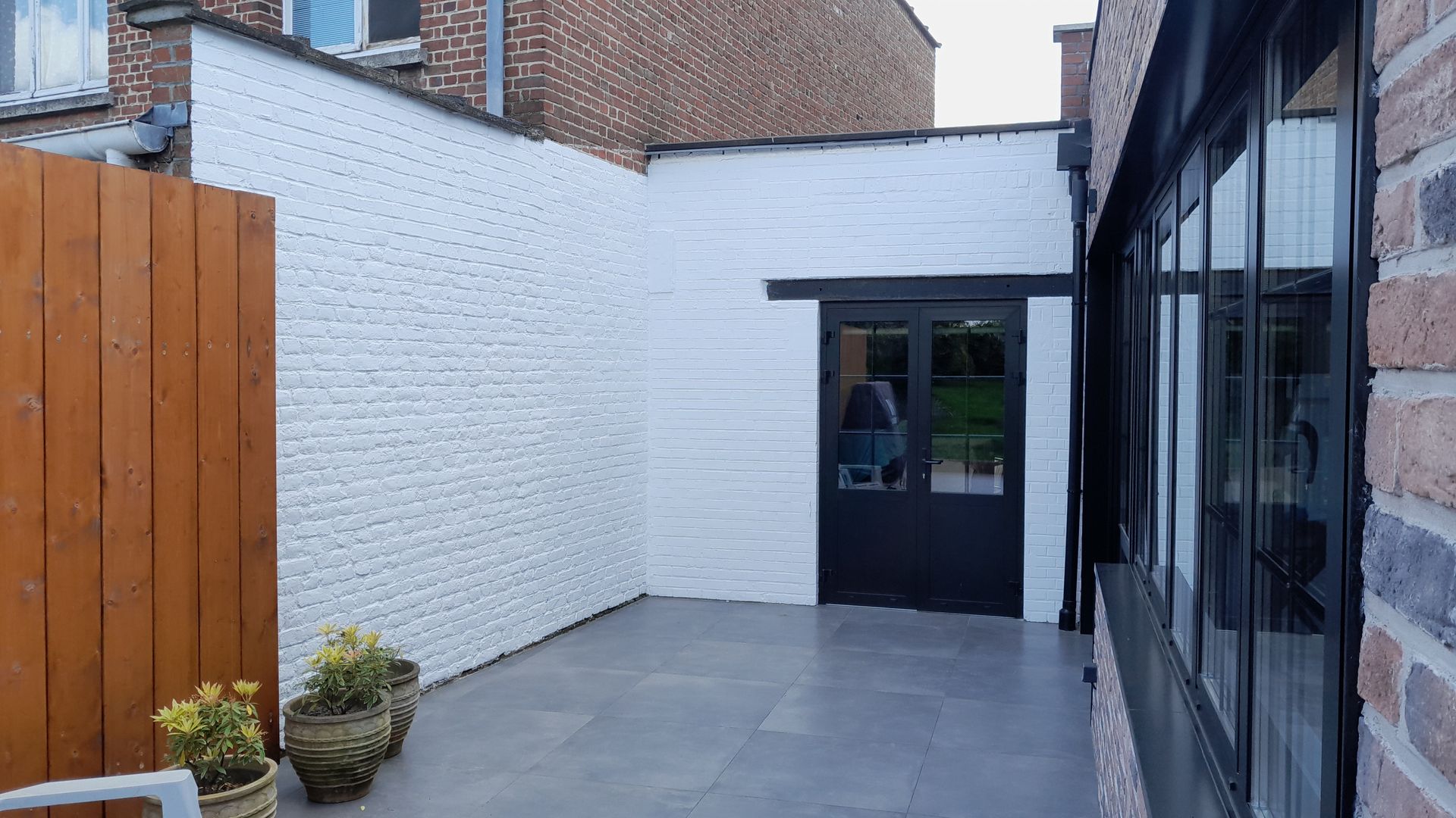 Cour extérieure avec murs en briques blanches peintes, porte sombre et sol en pierre grise. Clôture en bois et plantes sur la gauche.