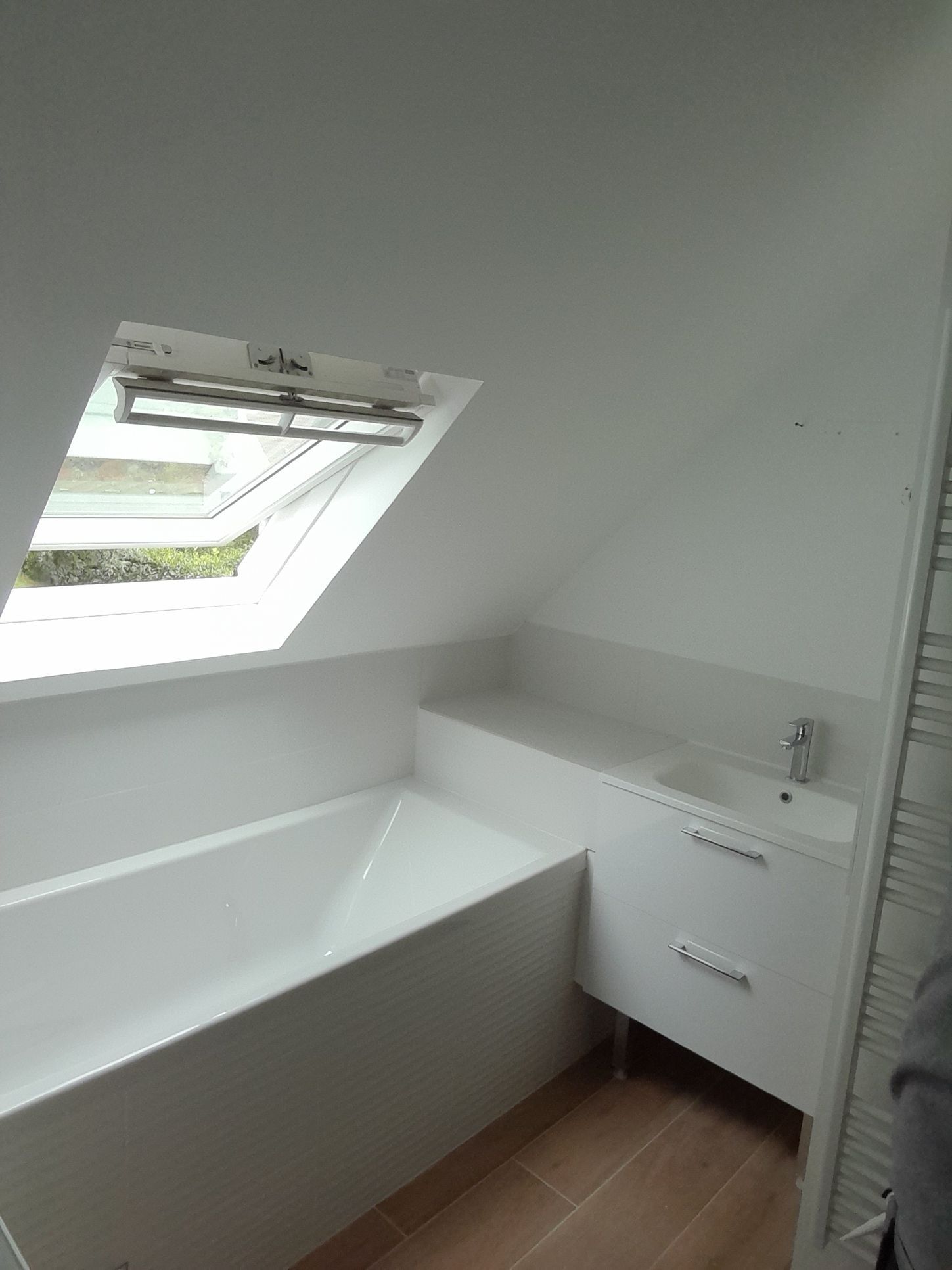 Salle de bains avec baignoire blanche, lavabo et puits de lumière dans un plafond mansardé.