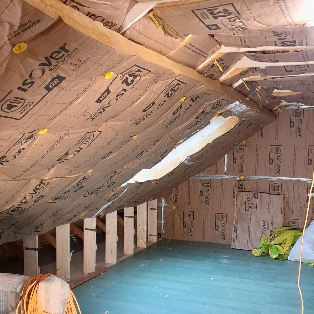 Intérieur d'un grenier en cours d'aménagement ; isolation installée au plafond et aux murs. Sol vert, poutres en bois.