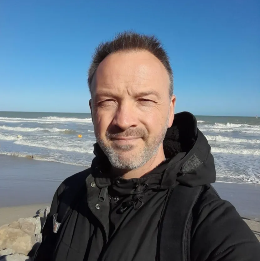 Un homme aux cheveux courts et vêtu d'une veste noire sourit sur une plage, l'océan en arrière-plan.