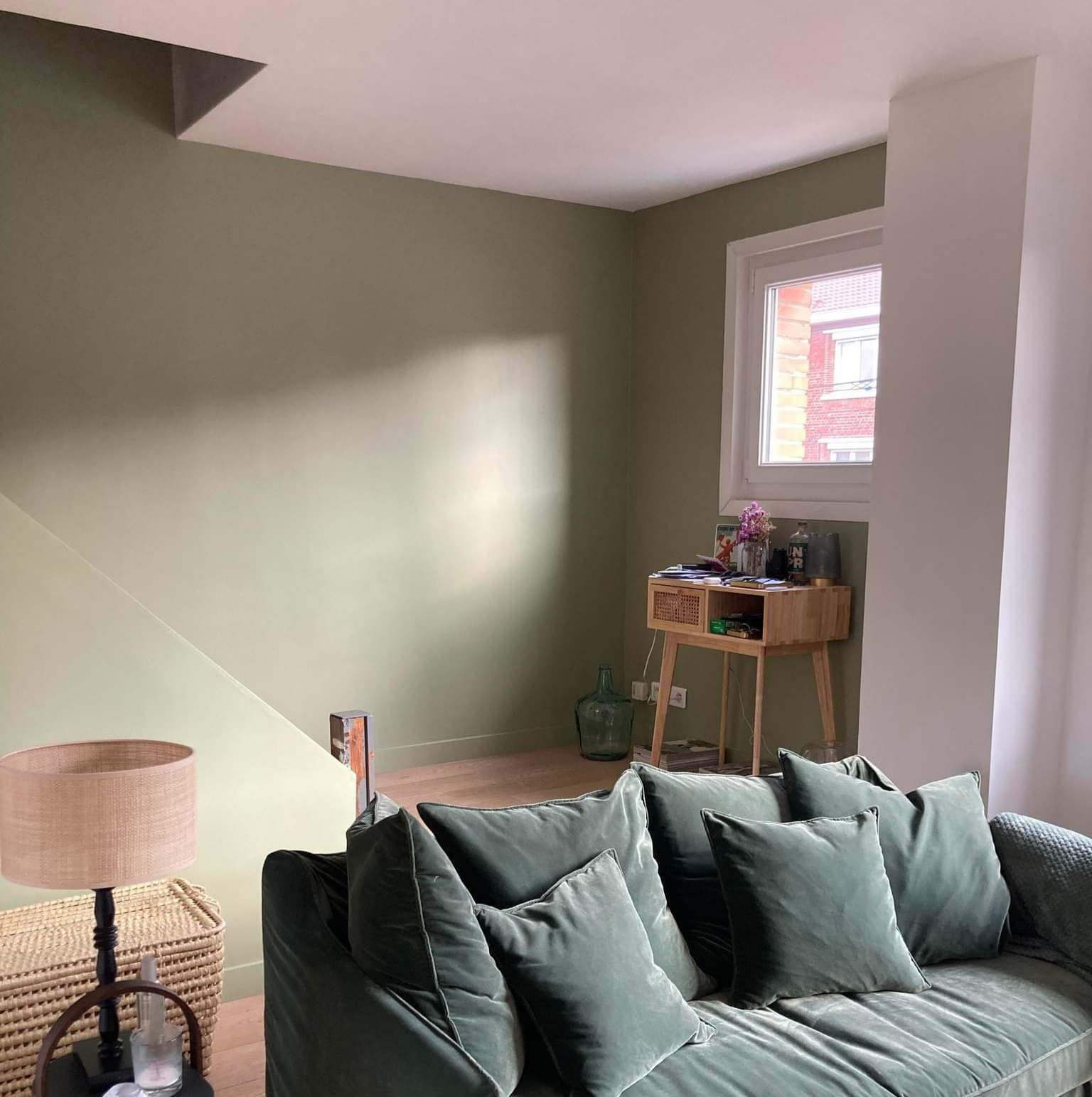 Chambre confortable avec murs verts et canapé, lumière naturelle par la fenêtre, table d'appoint en bois.