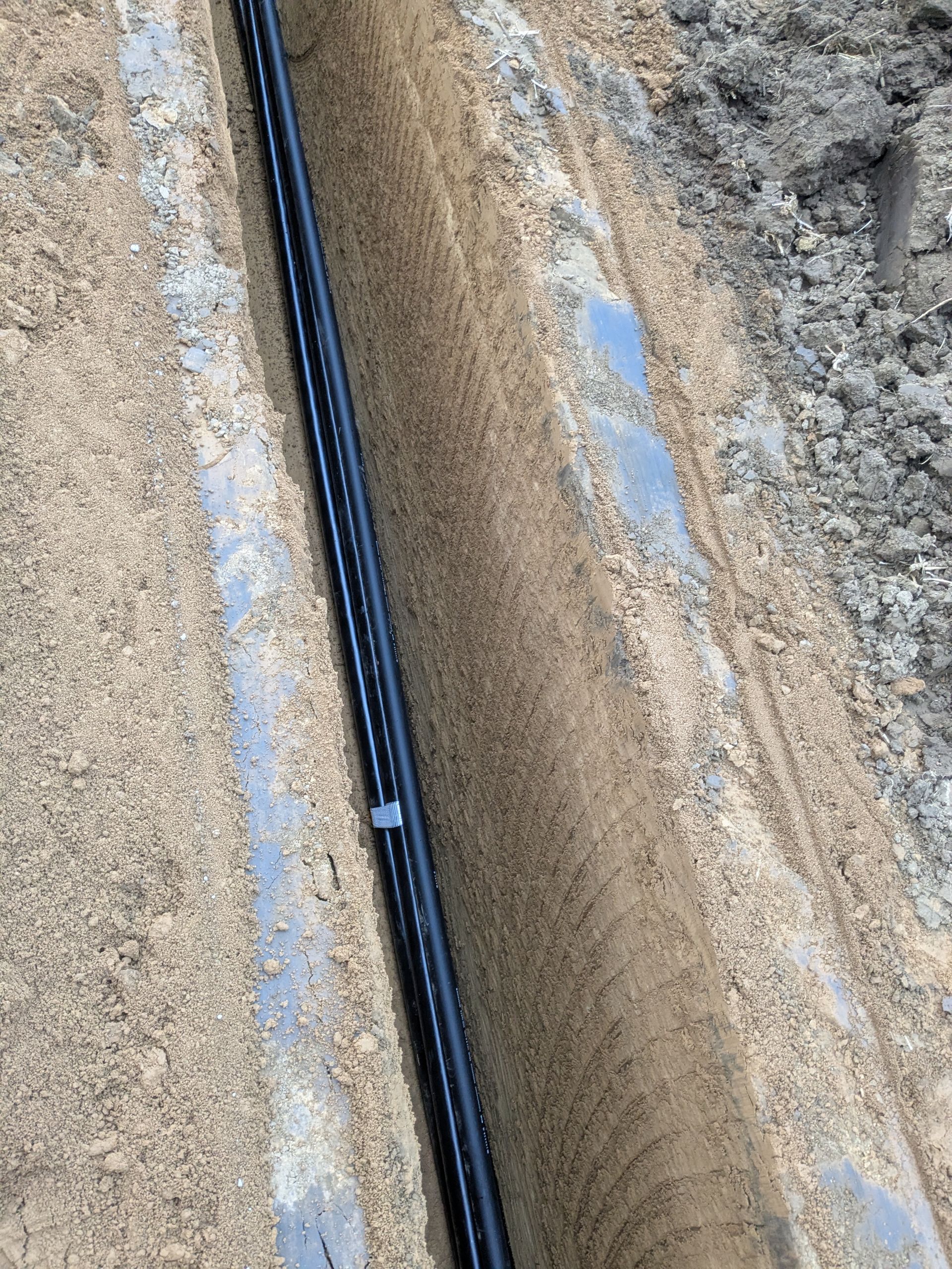 Ein schwarzes Rohr liegt im Dreck neben einer Betonmauer.