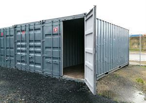 Ein grauer Frachtcontainer mit offener Tür steht auf einem Feld aus Erde.
