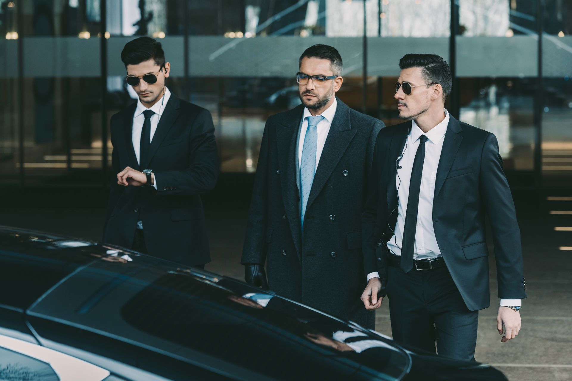 Trois hommes en costume portant des lunettes de soleil marchent à côté d'une voiture noire devant un bâtiment moderne.
