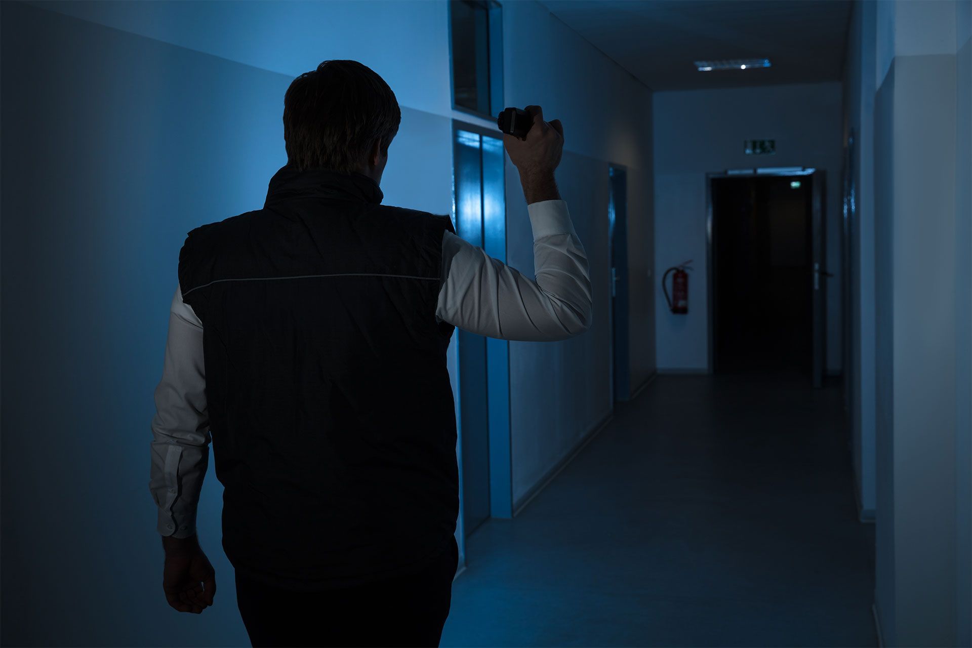 Une personne dans un couloir sombre, vue de dos, tenant une lampe torche et regardant vers une porte sombre.