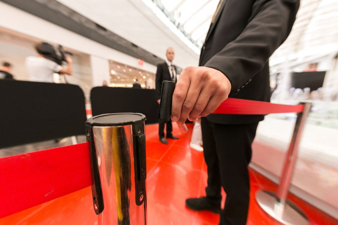 Une personne en costume se tient près d'une corde de poteau rouge dans un hall moderne et lumineux, avec des photographes en arrière-plan.