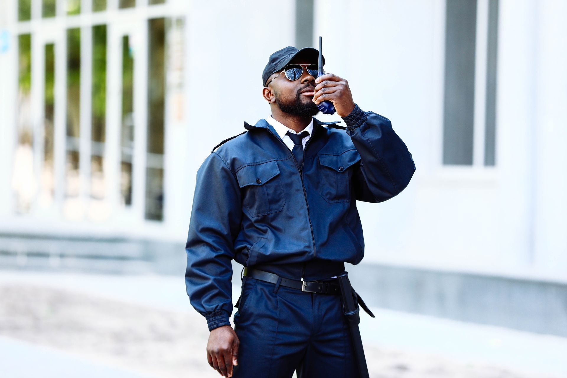 Un agent de sécurité en uniforme bleu foncé et lunettes de soleil parle dans un talkie-walkie à l'extérieur d'un bâtiment.
