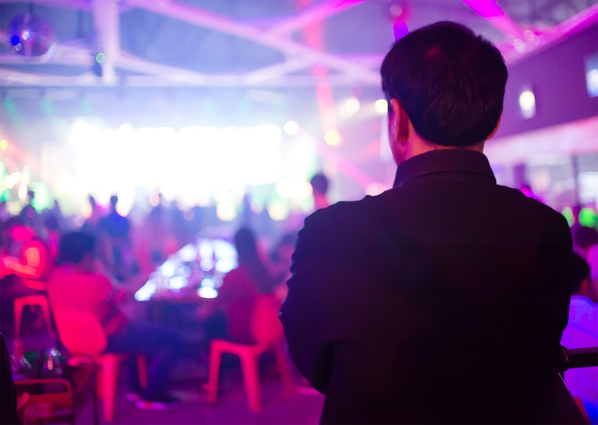 Vue de dos d'une personne portant une veste sombre regardant une boîte de nuit bondée et faiblement éclairée, avec des projecteurs de scène aux couleurs rose et bleu éclatantes.