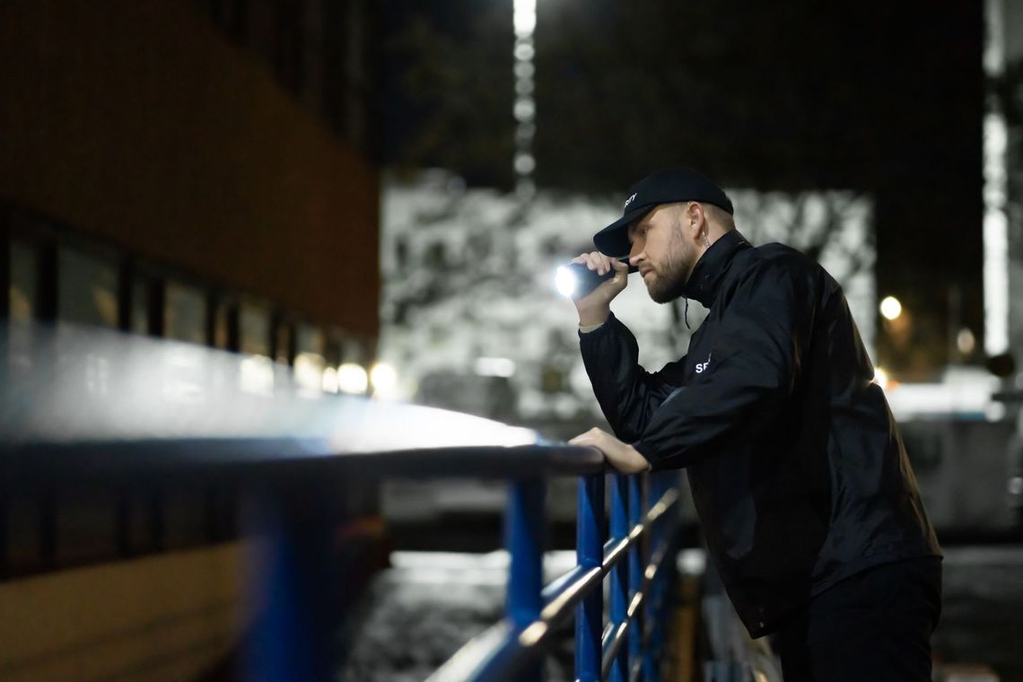 Une personne vêtue d'une veste sombre et d'une casquette de baseball s'appuie sur une rambarde bleue, la nuit, et éclaire le loin avec une lampe torche.