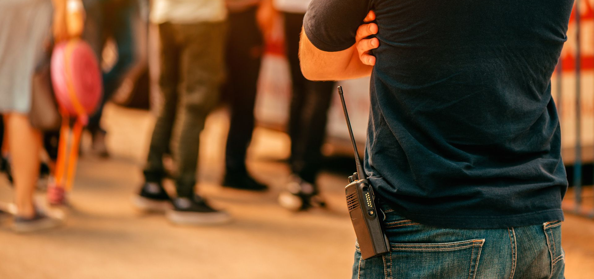 Un agent de sécurité, les bras croisés et une radio accrochée à la ceinture, se tient près d'un groupe de personnes à l'extérieur.