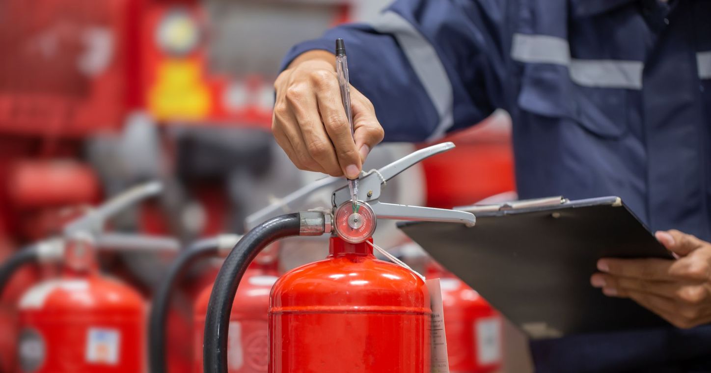 Une personne en uniforme bleu inspecte un extincteur rouge tout en tenant un bloc-notes et un stylo.