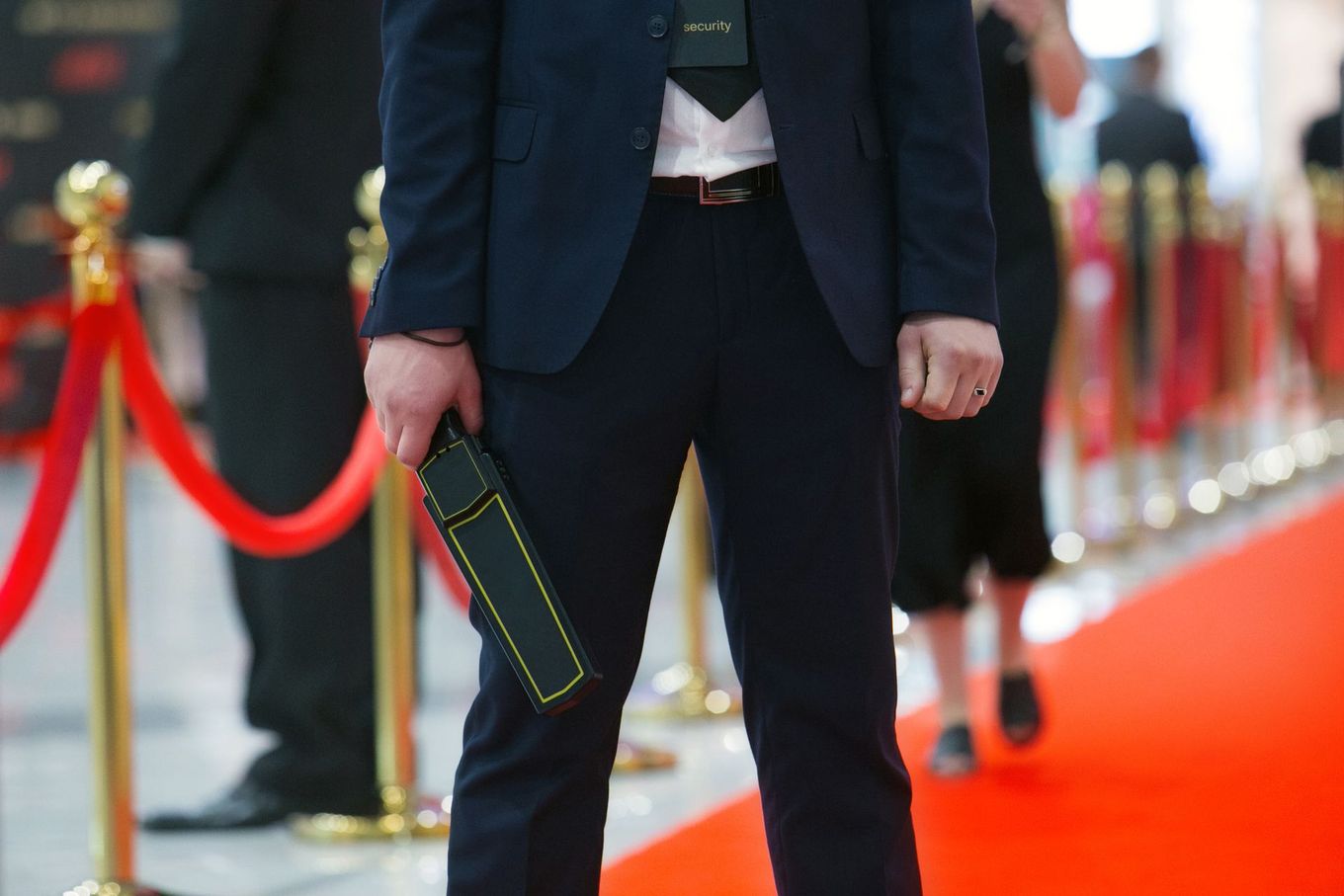 Un agent de sécurité en costume se tient sur un tapis rouge bordé de poteaux, tenant un détecteur de métaux portatif.