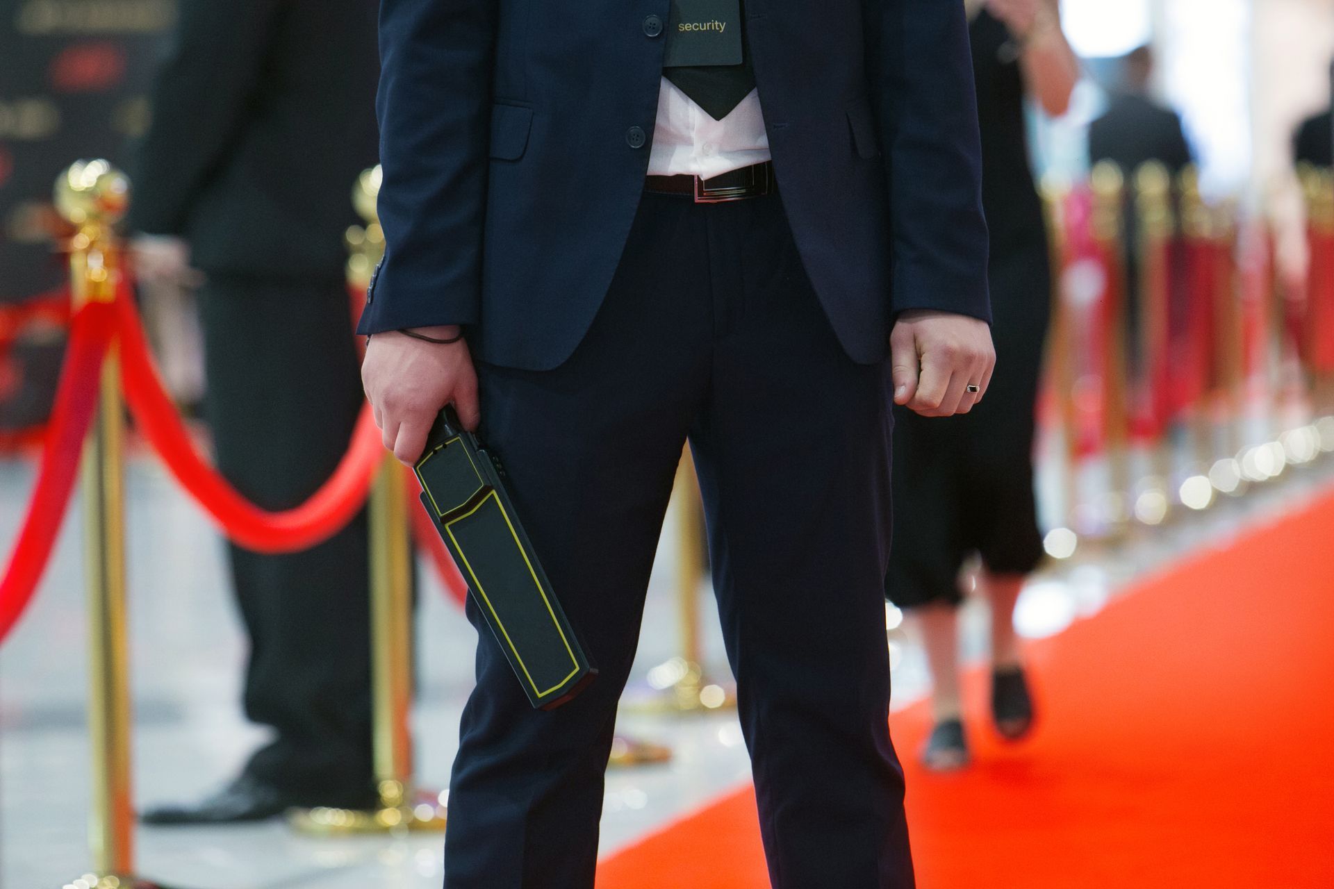 Un agent de sécurité en costume se tient sur un tapis rouge bordé de poteaux, tenant un détecteur de métaux portatif.