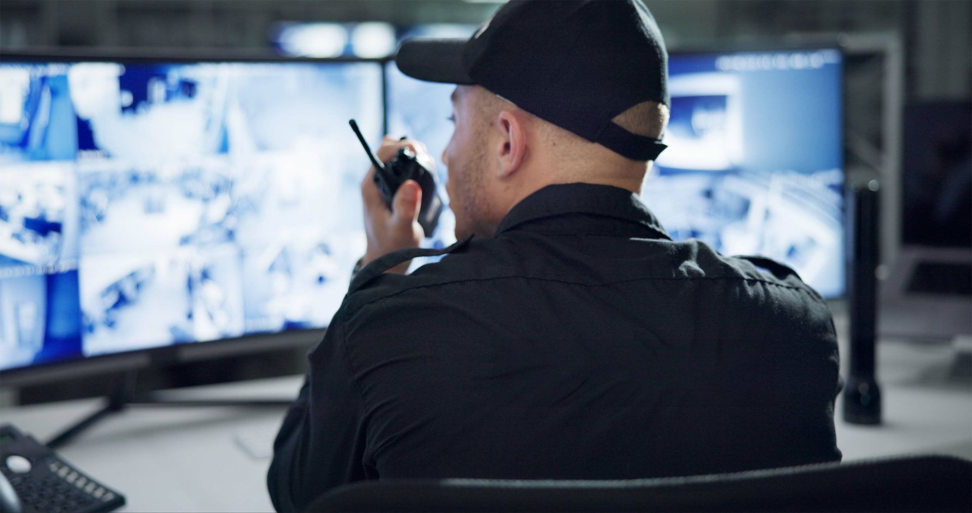 Un agent de sécurité en uniforme et casquette noirs est assis dans un bureau sombre, parlant dans un talkie-walkie tout en regardant des écrans.