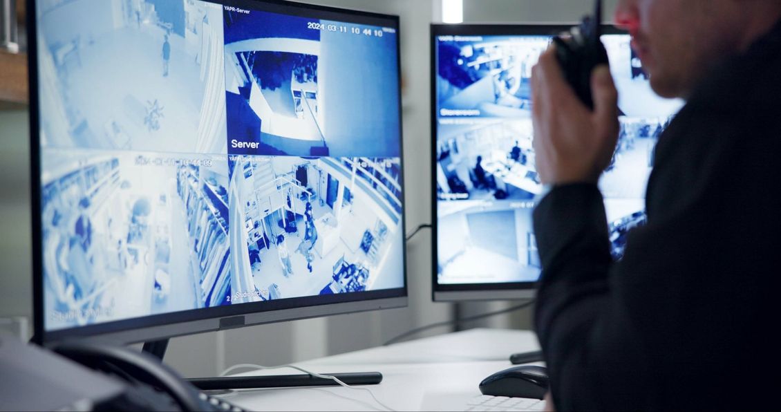 Un agent de sécurité surveille les flux de plusieurs caméras de surveillance sur deux écrans d'ordinateur tout en tenant une radio.