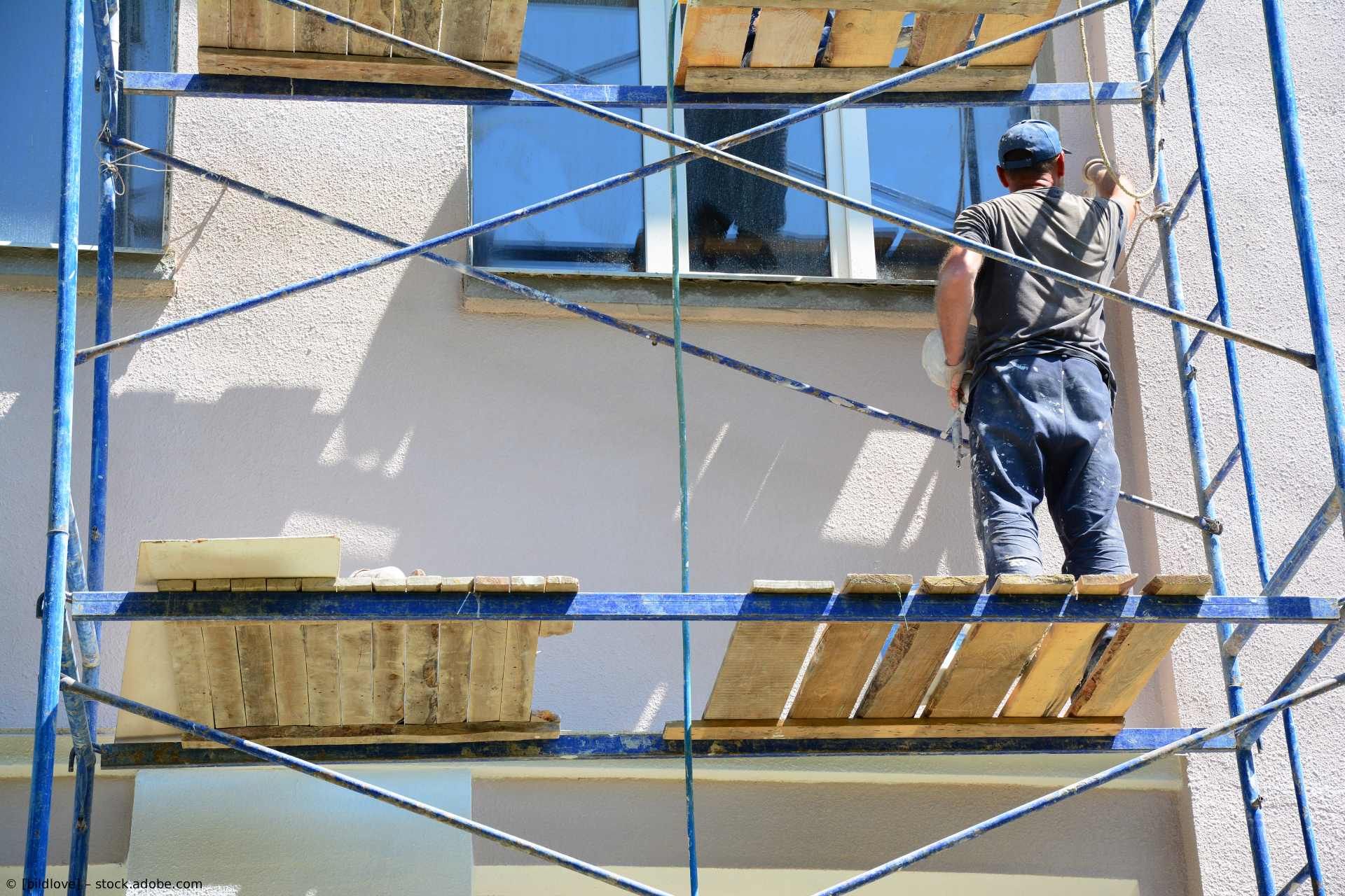 Eine Person steht auf einem Metallgerüst und streicht die Außenwand eines Gebäudes in der Nähe eines Fensters.