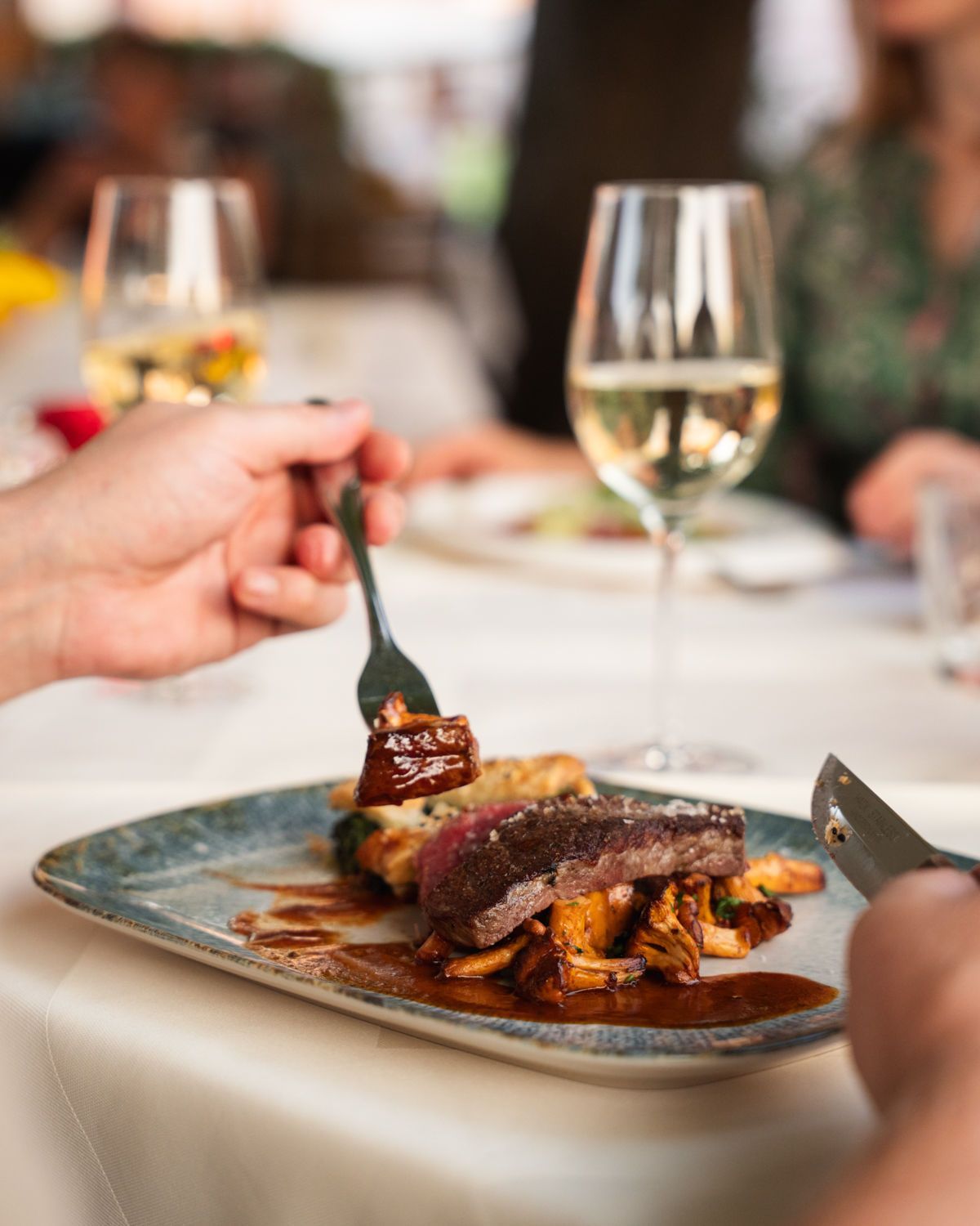 Ein auf einem Restauranttisch angerichtetes Steakgericht, das neben einem Glas Weißwein mit Messer und Gabel zerteilt wird.