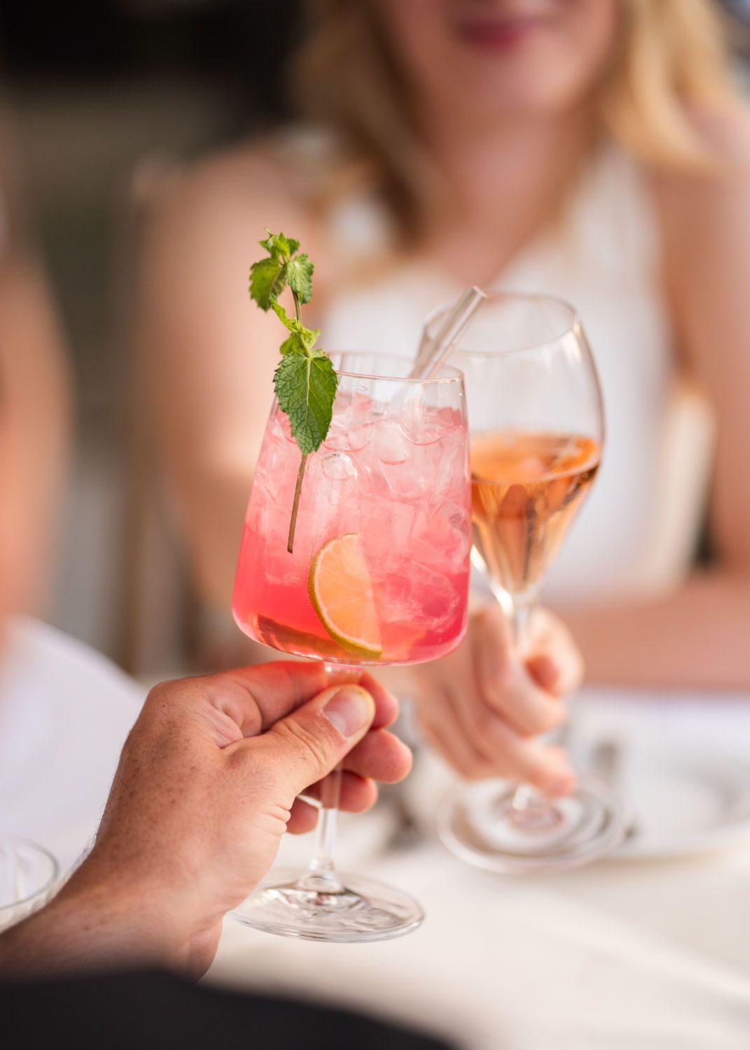 Eine Hand hält einen rosafarbenen Cocktail mit Minzgarnitur neben einem Glas Roséwein auf einem Tisch.