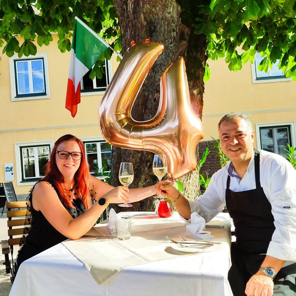 Paar stößt mit Wein an einem Tisch im Freien an, goldener Ballon mit der Zahl „4“, italienische Flagge im Hintergrund.