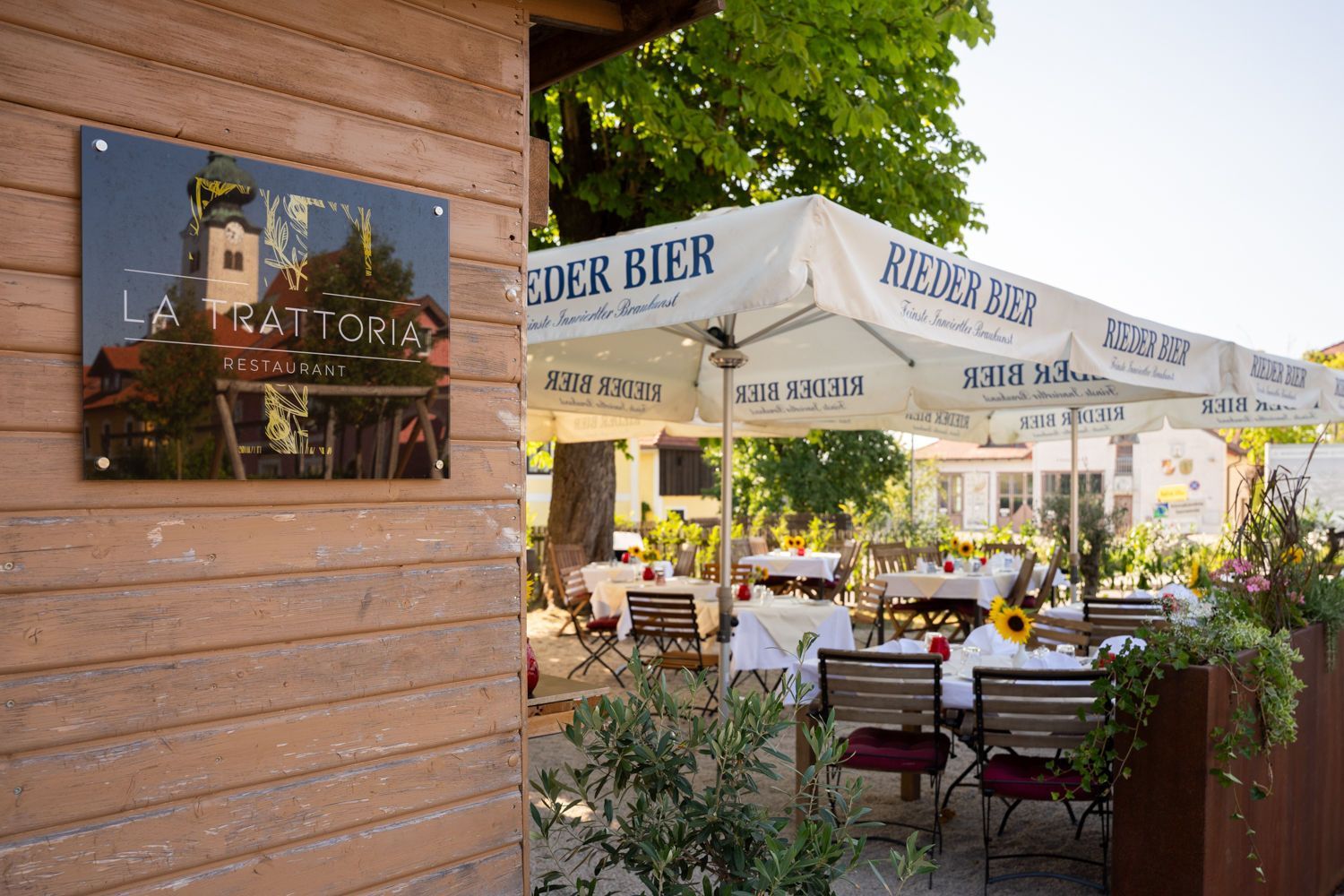 Terrasse eines Restaurants im Freien mit weißen Sonnenschirmen, Tischen und Blumen neben einem hölzernen Wandschild.