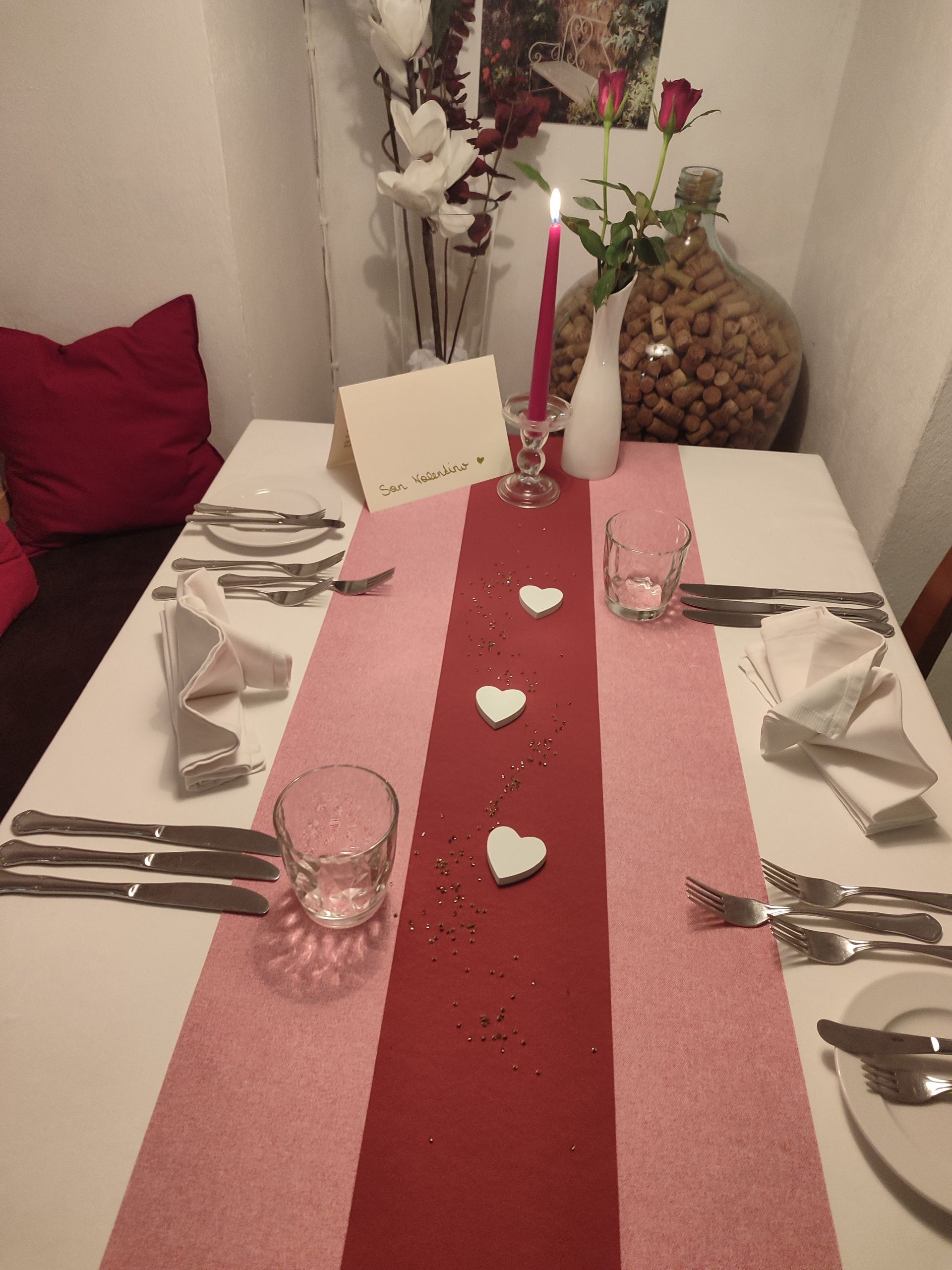 Valentinstagstisch für zwei Personen mit rosa und roten Dekorationen, Kerzen und weißen Herzen.