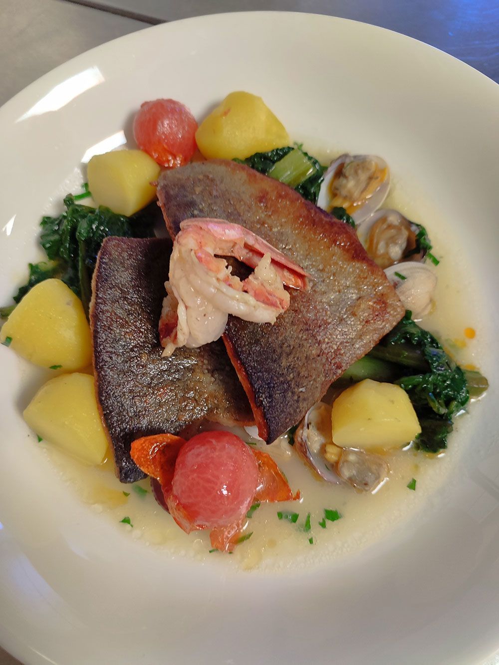 Gebratener Fisch mit Gemüse, Kartoffeln, Muscheln und Garnelen auf einem weißen Teller.