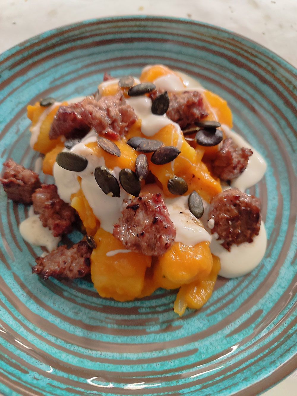 Orangefarbene Gnocchi mit Wurst, weißer Soße und Kürbiskernen auf einem türkisfarbenen Teller.