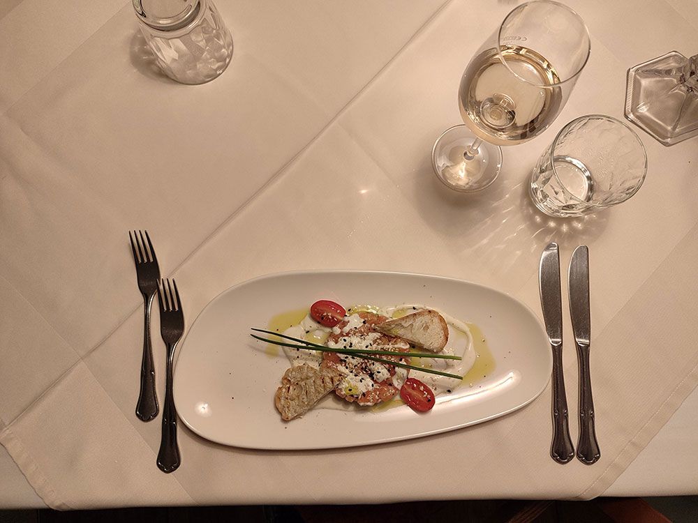 Draufsicht auf ein Tellergericht mit Tomaten und Brot, serviert auf einer weißen Tischdecke, mit Besteck und Getränken.