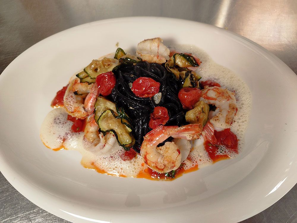 Schwarzes Nudelgericht mit Garnelen, Tomaten, Zucchini und weißem Schaum auf einem weißen Teller.