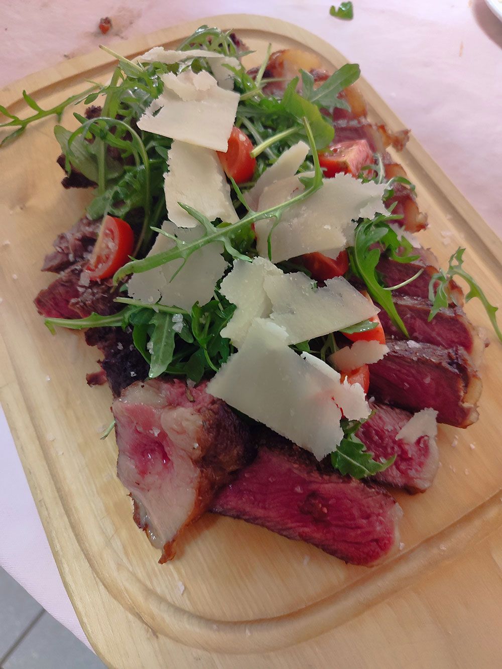 Geschnittenes Steak mit Rucola, Kirschtomaten und gehobeltem Parmesan auf einem Holzbrett.