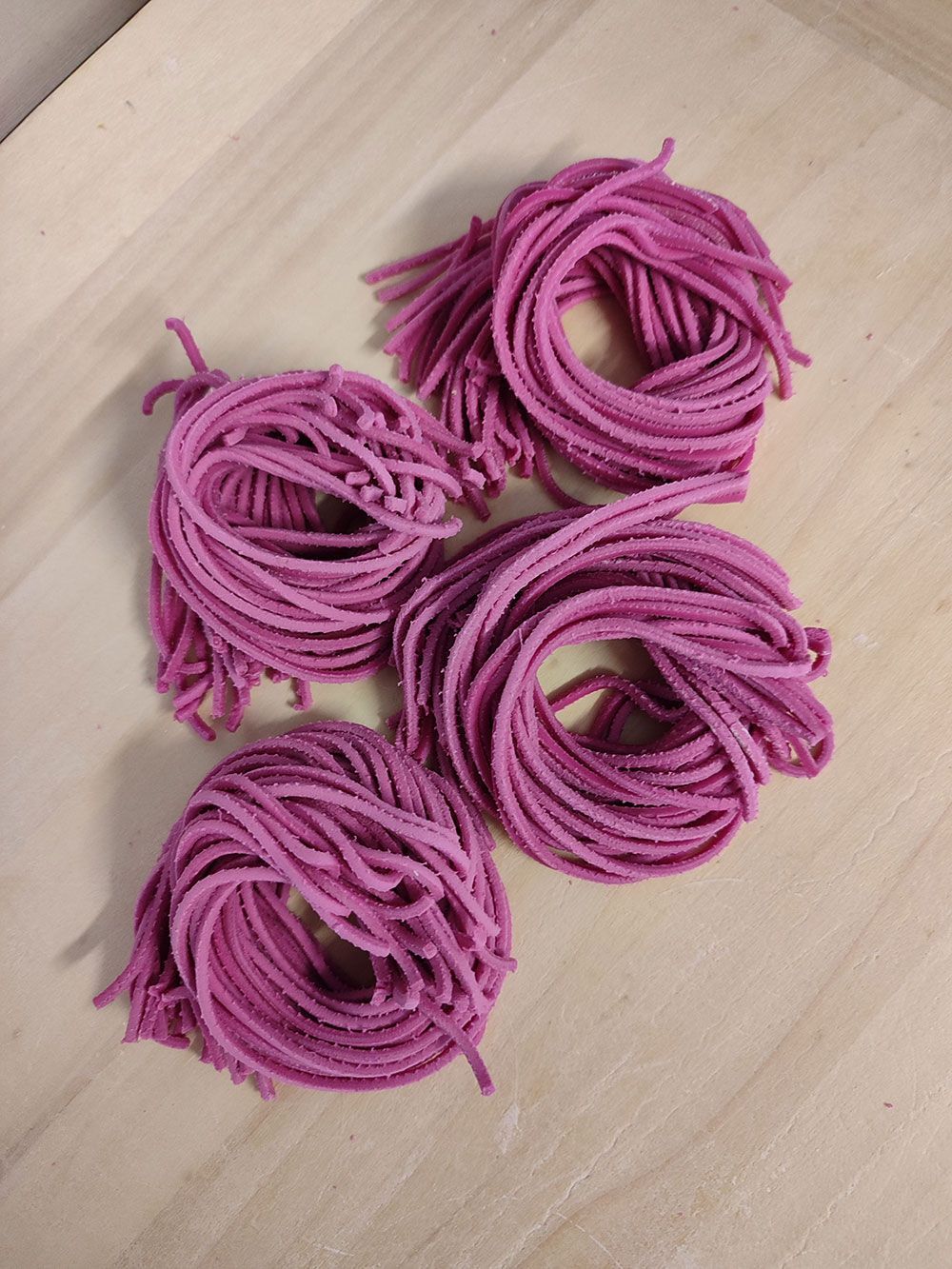 Vier runde Nester aus leuchtend rosa Spaghettinudeln auf einer hellen Holzoberfläche.