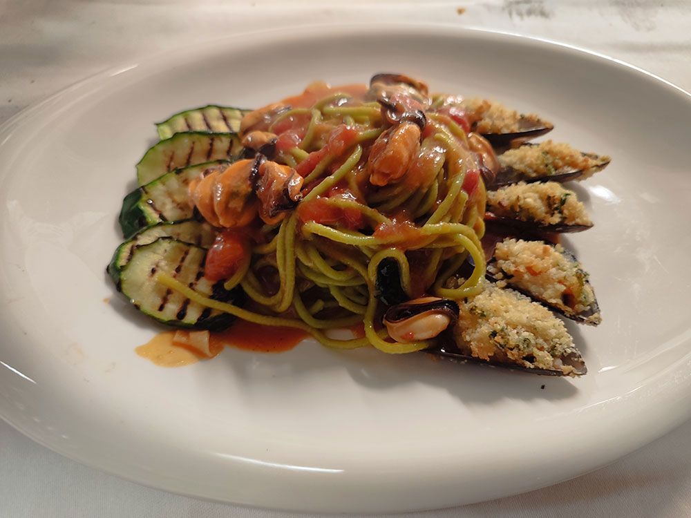 Nudelgericht: Grüne Nudeln mit Tomatensoße, gegrillte Zucchini und gebackene Muscheln auf einem weißen Teller.