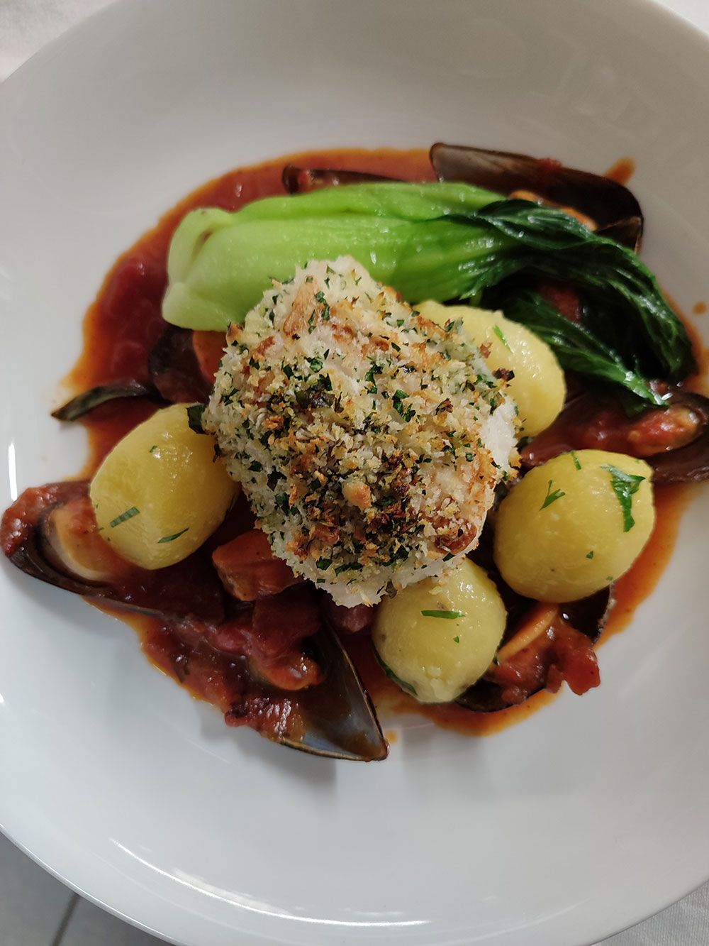 Tellergericht: Fisch mit Panade, Kartoffeln, Pak Choi und Muscheln in roter Sauce.