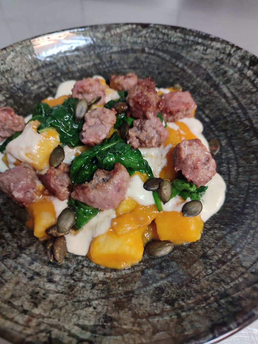 Gnocchi-Gericht mit Wurst, Spinat, Kürbiskernen und Sahnesauce in einer grau gesprenkelten Schüssel.