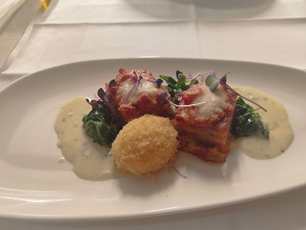 Weißer Teller mit zwei mit Fleisch belegten Stapeln, frittierten Bällchen, grünem Gemüse und Sahnesoße.