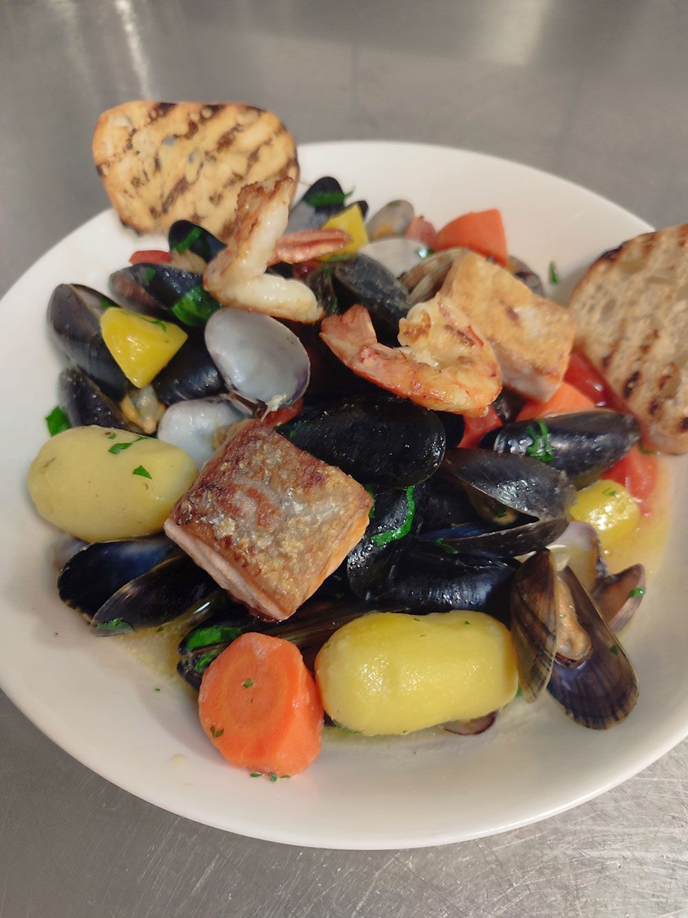 Meeresfrüchteeintopf in einer weißen Schüssel mit Muscheln, Garnelen, Kartoffeln, Karotten und gegrilltem Brot.